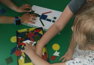 Dzieci przy stoliku układają z mozaiki geometrycznej robota według instrukcji